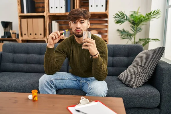 Arab man with beard working on depression holding pills and water skeptic and nervous, frowning upset because of problem. negative person.