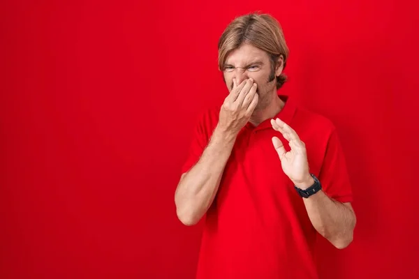 一个白种人 留着胡子 站在红色的背景上 闻到一股难闻 无法忍受的气味 用手指捂住鼻子呼吸 — 图库照片