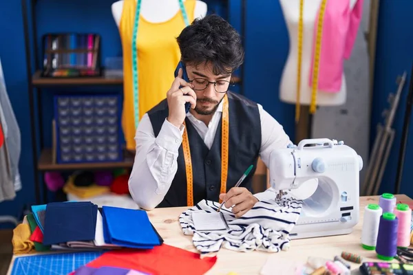 Young Hispanic Man Tailor Talking Smartphone Writing Notebook Sewing Studio — Stock Fotó