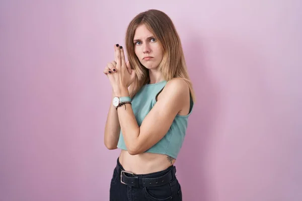 Blonde caucasian woman standing over pink background holding symbolic gun with hand gesture, playing killing shooting weapons, angry face