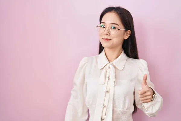 若い中国人女性がピンクの背景に立って手で幸せな親指をジェスチャーアップを行う 成功を示すカメラを見ると — ストック写真