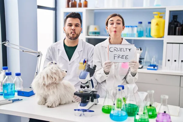 Young hispanic people working at scientist laboratory with dog making fish face with mouth and squinting eyes, crazy and comical.