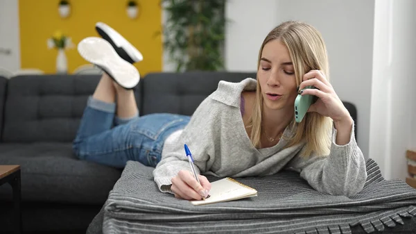 在笔记本上写字的金发年轻女人躺在沙发上 在家里讲电话 — 图库照片
