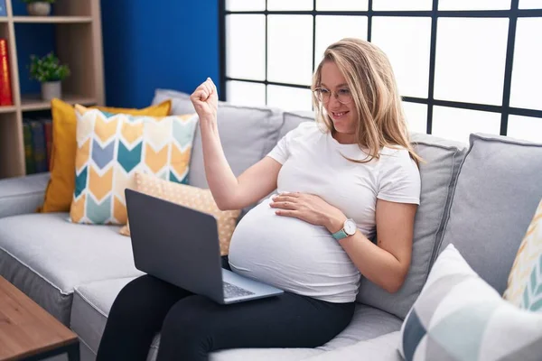 年轻的孕妇坐在沙发上 用笔记本电脑高喊着自豪 高举双臂庆祝胜利和成功 — 图库照片