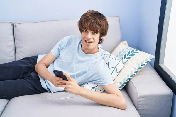 若いですブロンド男使用してスマートフォン嘘上のソファにホーム — ストック写真