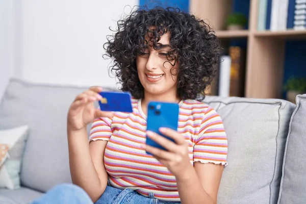 年轻的中东女人坐在家里沙发上 用智能手机和信用卡 — 图库照片