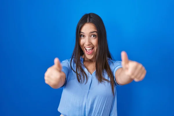 年轻的黑发女人站在蓝色的背景上 赞成用手做积极的手势 微笑着竖起大拇指 为成功感到高兴 优胜手势 — 图库照片
