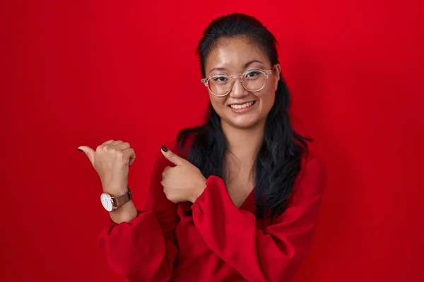 亚洲的年轻女子站在红色的背景上 双手和大拇指朝后 自信地微笑着 — 图库照片