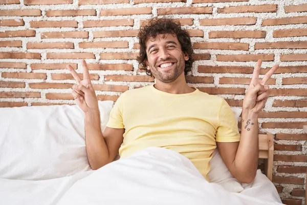 Young Hispanic Man Doing Victory Gesture Sitting Bed Bedroom — Zdjęcie stockowe