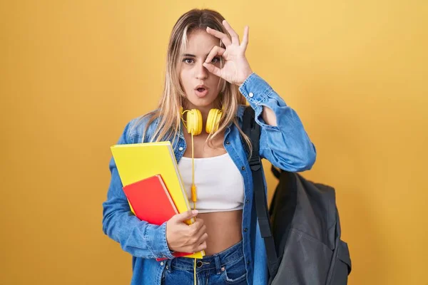 若いブロンドの女性学生のバックパックを着用し 本を保持本のジェスチャーを行う驚きの顔でショックを受け 目の指を見て 信じられない表現 — ストック写真
