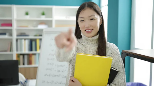 Young chinese woman student holding books pointing with finger to camera at library university