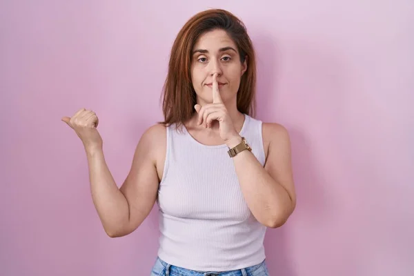 ピンクの背景に立っているブルネットの女性は 唇に手で側に指して指で静かになるように求めている 沈黙と秘密の概念 — ストック写真