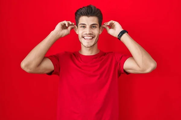 Kırmızı Arka Planda Dikilen Genç Spanyol Adam Gülümsüyor Parmaklarıyla Kulaklarını — Stok fotoğraf