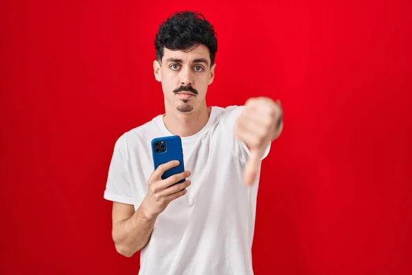 Латиноамериканец Использующий Смартфон Красном Фоне Выглядит Несчастным Злым Показывая Отказ — стоковое фото