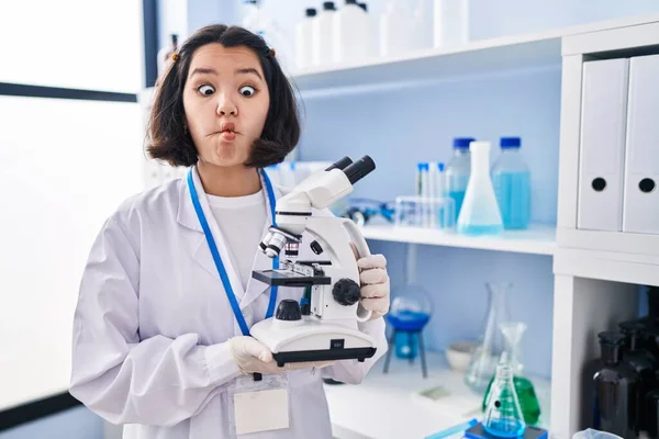 Young hispanic woman working at scientist laboratory holding microscope making fish face with mouth and squinting eyes, crazy and comical.