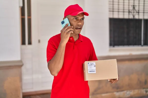 Young Latin Man Delivery Worker Holding Package Talking Smartphone Street — Stok fotoğraf