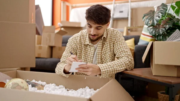 Young Arab Man Unpacking Cardboard Box Writing Notebook New Home — Stock Photo, Image
