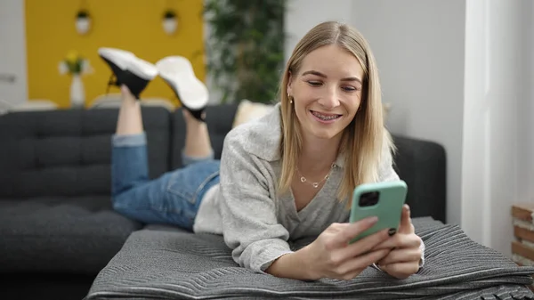 年轻的金发女人用智能手机躺在家里的沙发上 — 图库照片