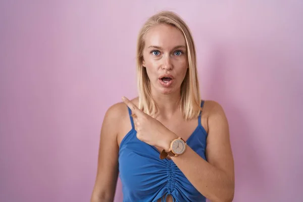 stock image Young caucasian woman standing over pink background surprised pointing with finger to the side, open mouth amazed expression. 