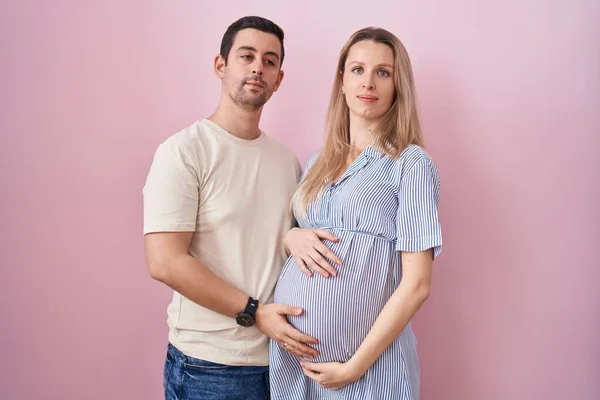 真面目な表情でリラックスしたピンクの背景の上に立つ赤ちゃんを期待している若いカップル カメラを見ているだけで — ストック写真