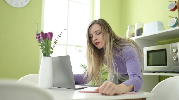 年轻的金发女人在家里用笔记本电脑在笔记本上写字 — 图库照片