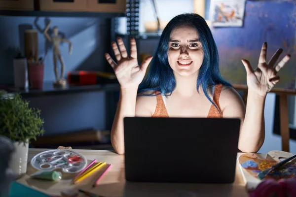 年轻的现代女孩 留着蓝色的头发 晚上带着笔记本电脑坐在艺术工作室里 用9号手指指指着 面带微笑 自信而快乐 — 图库照片