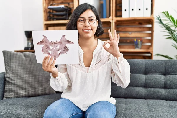 stock image Young hispanic therapist woman holding rorschach test doing ok sign with fingers, smiling friendly gesturing excellent symbol 