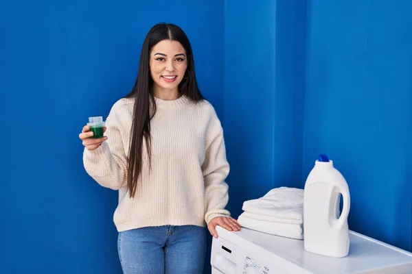 年轻美丽的惊慌失措的女人在洗衣房自信地拿着洗涤剂微笑 — 图库照片
