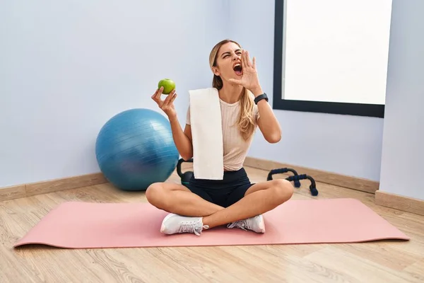 健康的なリンゴの叫びを保持し 口の上に手で横に大声で叫ぶスポーツウェアとタオルを身に着けている若いブロンドの女性 コミュニケーションの概念 — ストック写真