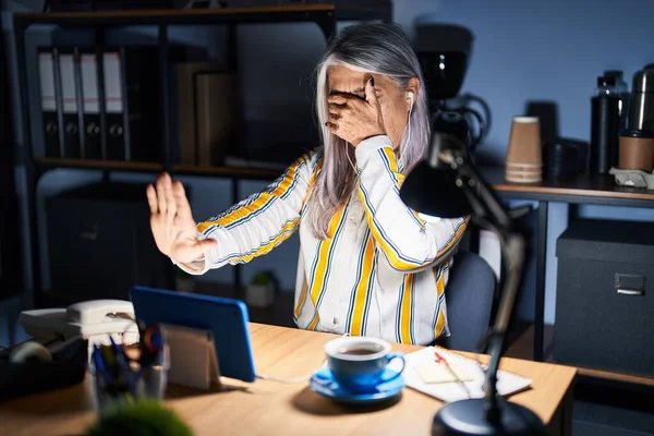 深夜オフィスで働く白髪の中年女性が手で目を覆い悲しみと恐怖の表情でストップジェスチャー 恥をかかせ否定的な概念 — ストック写真