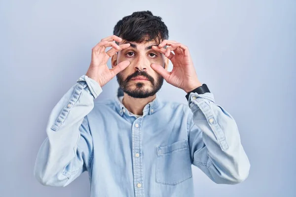 Молодой Латиноамериканец Бородой Стоит Синем Фоне Пытаясь Открыть Глаза Пальцами — стоковое фото