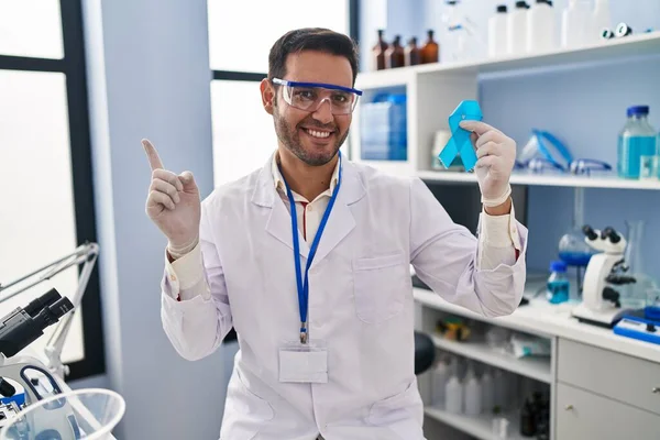 Młody Latynos Brodą Pracujący Laboratorium Trzymający Niebieską Wstążkę Uśmiechający Się — Zdjęcie stockowe