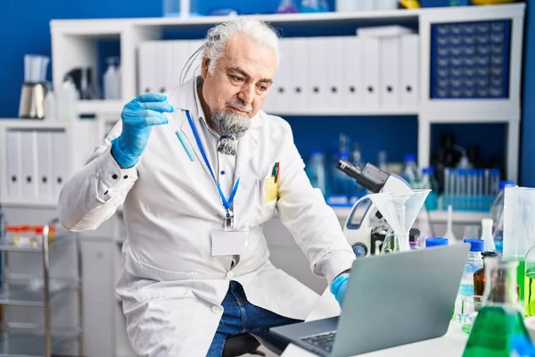 Middle age grey-haired man scientist using laptop holding test tube at laboratory