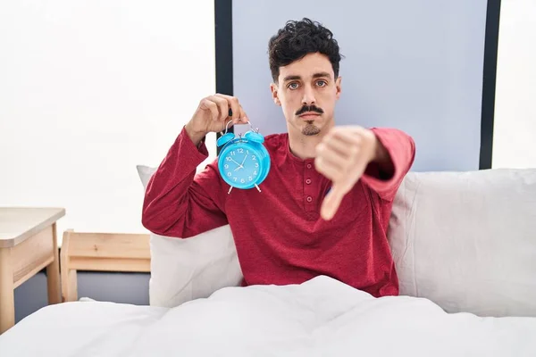 Hispanic Man Håller Väckarklocka Sängen Med Argt Ansikte Negativa Tecken — Stockfoto