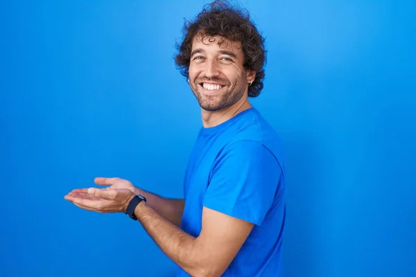 西班牙裔年轻人站在蓝色的背景上 手拉手张开手掌 展示复制的空间 展示广告时面带微笑 喜形于色 — 图库照片