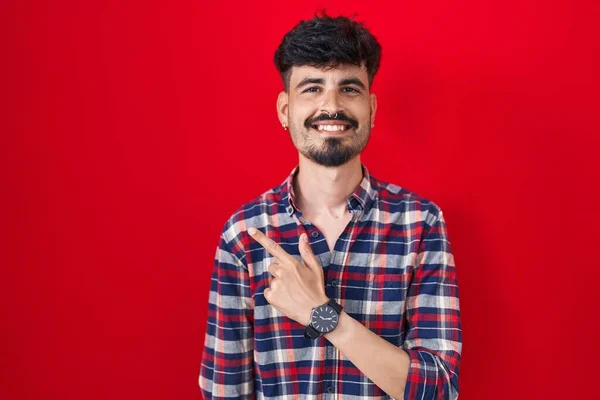 髭を生やした若いヒスパニック系の男が笑顔で陽気に赤い背景の上に立ち 幸せと自然な表情で側に手と指を指しています — ストック写真