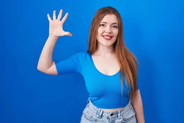 自信と幸せ笑いながら指の数5を見せ 指差す青い背景に立って赤毛の女性 — ストック写真