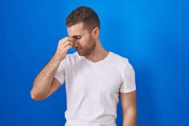 Mavi arka planda dikilen genç beyaz adam yorgun burnu ve gözleri ovuşturup yorgun ve baş ağrısı çekiyor. Stres ve hüsran kavramı. 