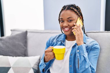 Afro-Amerikalı kadın akıllı telefondan konuşuyor evde kahve içiyor.