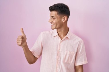 Young hispanic man standing over pink background looking proud, smiling doing thumbs up gesture to the side 