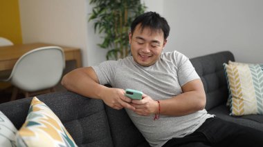 Young chinese man using smartphone sitting on sofa at home