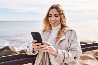 Young blonde woman smiling confident using smartphone at seaside