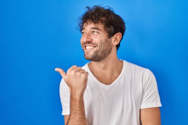 Hispanic young man standing over blue background smiling with happy face looking and pointing to the side with thumb up. 