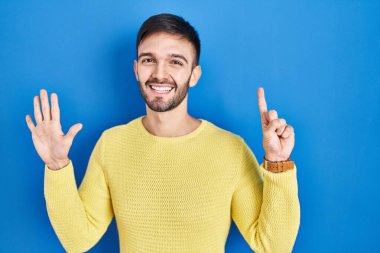 Hispanic man standing over blue background showing and pointing up with fingers number six while smiling confident and happy. 