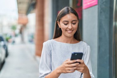 Genç, güzel, İspanyol bir kadın sokakta akıllı telefon kullanarak gülümsüyor.