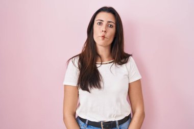 Young brunette woman standing over pink background making fish face with lips, crazy and comical gesture. funny expression. 
