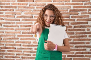 Elinde sanat defteri tutan beyaz bir kadın seni ve kamerayı işaret ediyor, gülümsüyor olumlu ve neşeli. 