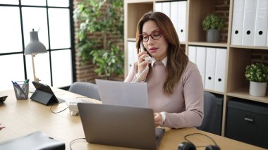 Young beautiful hispanic woman business worker talking on telephone reading document at office