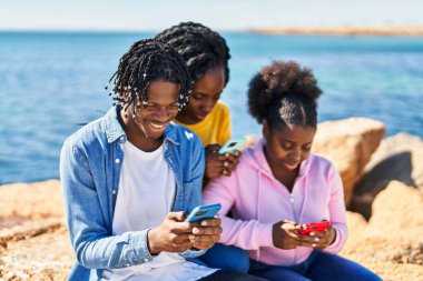 African american friends using smartphone sitting on rock at seaside