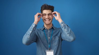 Genç İspanyol adam gülümsüyor. Gözlük takıyor, mavi arka planda.